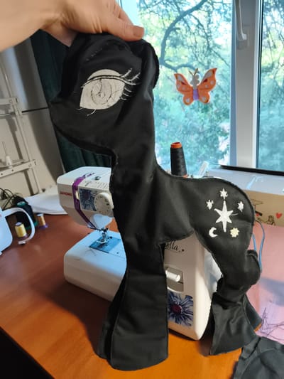 A person's hand is holding a black stuffed toy, possibly a horse or pony, in front of a white Janome sewing machine. The toy has an embroidered eye and a constellation of stars and a crescent moon on its hindquarters. The sewing machine is on a wooden desk next to a window with a view of trees and a small orange butterfly decoration. Spools of thread are visible on the desk and near the sewing machine. The scene appears to be indoors, likely a home craft room, in Sokolivske, Ukraine.