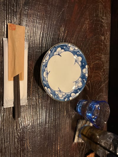 The image shows a wooden table with a small blue and white patterned plate, a pair of chopsticks wrapped in paper and a white napkin, and two glasses. One glass is filled with water and the other with a dark liquid. There are no people in the image. The location is the Benesse House Museum on Naoshima Island, Japan, with coordinates 34.458, 133.995. The wooden table is dark brown and appears to be made of planks of wood. The table is scratched and worn. The chopsticks are dark brown and appear to be wooden. The napkin is white and has a plastic wrapper. The blue glass has a blue tint and appears to be frosted. The glass with the dark liquid has a brown tint and has a black straw. The plate is empty and is a decorative saucer with a blue and white flower pattern. The scene is a typical dining arrangement at the Benesse House Museum. The presence of chopsticks and the patterned plate imply a setting in a Japanese restaurant.