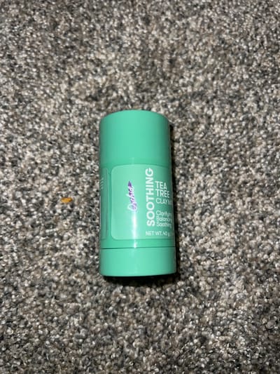 A green cylindrical stick of Orchid "Soothing Tea Tree Clay Mask" lies on a grey, textured carpet. The label on the stick is white and green with purple accents, and it reads "Orchid," "SOOTHING," "TEA TREE," "CLAY MASK," "Clarifying," "Balancing," "Soothing," and "NET WT. 40 g | 1.4 oz." The product is positioned vertically on the carpet, with the top of the lid not visible in the frame. The carpet appears to be in an indoor setting. There is no information in the image to determine the location in Everett, United States of America.