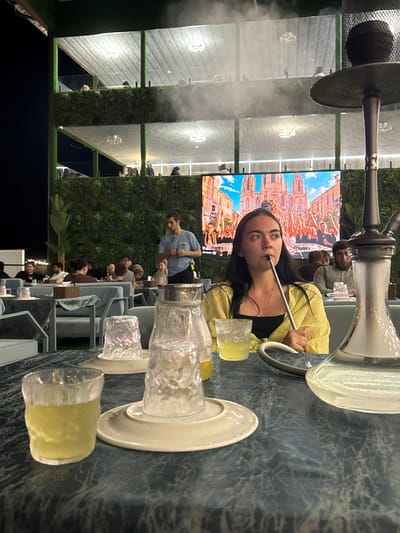 In Derbent, Russia, a woman is seated at a table, holding a shisha hose, and appears to be smoking. She is wearing a yellow shirt and a black top. In front of her are several glasses and a shisha pipe. The table is covered with a patterned cloth. In the background, a large screen displays a vibrant image of a crowd and a cathedral. Other patrons are visible in the background, seated at tables, and a waiter is standing. The environment appears to be an outdoor or semi-outdoor establishment with lush green walls and multi-level seating. The lighting suggests it is evening or night.