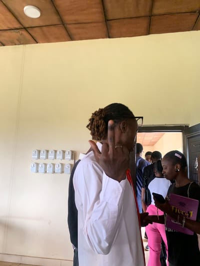 The image shows an indoor scene with several people. In the foreground, a person wearing a white collared shirt is facing away from the camera, with their right hand raised in a gesture. This person has dreadlocks and is wearing glasses and a ring on their finger. To the right, a woman in a black top is looking down at a purple folder in her hands, which appears to be a "Five in 1" book. She has a pink ribbon in her hair and is wearing earrings and bracelets. Behind them, several other people are visible, including some standing near an open doorway that leads into another room with a tiled ceiling. On the wall to the left, there is a row of twelve circular electrical switches. The walls are painted a light yellow color, and the floor appears to be tiled. The setting is an indoor area, possibly a school or community center, in Farin Yashe, Nigeria.