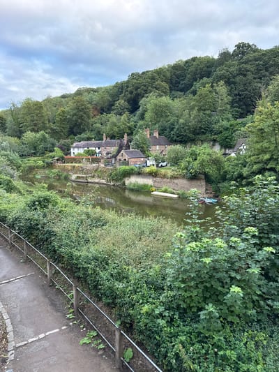 The image depicts a serene riverside scene in the town of Ironbridge, United Kingdom. A tranquil river flows through the center of the frame, reflecting the overcast sky and the lush greenery that surrounds it. On the riverbank, a cluster of quaint houses with traditional brick chimneys nestles amongst the trees. Several boats are moored along the water's edge, suggesting a popular spot for leisure activities.

To the left of the river, a paved pathway is bordered by a metal railing and dense vegetation, indicating a walking trail or promenade. The pathway leads the eye towards the houses and the natural beauty of the area. The overall impression is one of peaceful coexistence between human habitation and the natural environment, characteristic of the historic and picturesque town of Ironbridge.