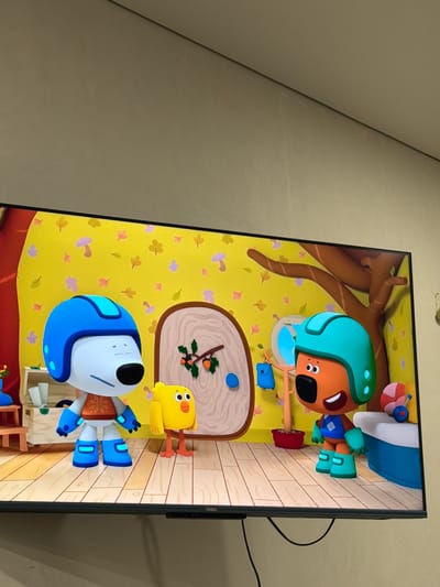 The media file shows a television screen displaying a cartoon scene. The scene features several animated characters, including a brown dog wearing a turquoise helmet and blue shorts, and a yellow bird with orange legs. Another character, a white robot with blue accents and a blue helmet, is also visible. The background is a bright yellow wall decorated with colorful autumn leaves and mushrooms. There is a wooden floor and a tree with brown branches. A small blue mouse and a red pot are also present. The cartoon appears to be aimed at children. The television set itself is black and has a "Haier" logo on the bottom right side. The overall mood of the scene is cheerful and playful. The image is taken in Krasnyi Krym, Russia. There is no discernible time of day or weather information, and no specific location cues beyond the country.
