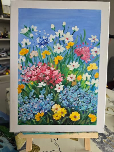 The image displays a vibrant, impasto acrylic painting of a wildflower bouquet against a light blue background. The flowers are rendered in a variety of colors including white, yellow, pink, and blue, with visible brushstrokes adding texture and depth. The bouquet is densely packed, with smaller blue flowers forming the base, transitioning to clusters of pink and red, and topped with scattered white and yellow blooms. Green stems and leaves are visible throughout the arrangement. The painting is positioned on a light-colored wooden easel, which is partially visible at the bottom of the frame. The background behind the easel is blurred and appears to be an indoor setting with furniture and shelves, suggesting the painting is displayed in a home or studio. There is no visible indication of the location being in Claremorris, Ireland, or any specific city.