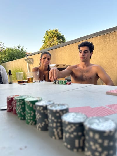 This is a medium shot, eye-level, outdoor photograph of a group of people playing poker at a table in a backyard in Zoran, Israel. The lighting suggests it is late afternoon or early evening, with the sun casting warm light on the scene. The background consists of a beige stucco wall, some green foliage, and a clear blue sky.

In the foreground, a white plastic table is set up with stacks of poker chips in red, green, and gray, as well as a deck of red-backed playing cards. The chips are arranged in a line, with the red chips closest to the viewer and the gray chips further away.

To the left of the table, a person wearing a white t-shirt is partially visible. Next to them, there is a bottle of spray and three plastic cups, two of which contain amber-colored beverages.

In the middle ground, a woman with long dark hair is seated at the table, looking towards the camera with a slight smile. She is wearing a patterned top. A man with dark curly hair, shirtless, is seated to her right, reaching out and touching a stack of poker chips. His body is angled away from the camera.

The overall atmosphere appears relaxed and social, with the group engaged in a game of poker in a pleasant outdoor setting.