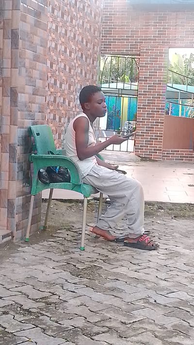 A young person, appearing to be of school age, sits on a green plastic chair. They are wearing a white tank top and light-colored pants, with dark sandals on their feet. The person is holding a mobile phone in their right hand and appears to be looking at it. Their left arm rests on the armrest of the chair. The chair is positioned on a paved outdoor area with a herringbone pattern of grey bricks. To the left, a wall is constructed from beige and brown tiles. In the background, a brick wall with windows and metal bars is visible, behind which greenery and possibly part of a building with a railing can be seen. The overall impression is a casual outdoor setting, possibly a residential area or a public space within the city of Agbarho, Nigeria.