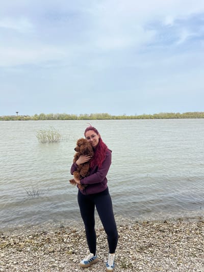 A woman with red hair stands on a gravel beach next to a body of water, holding a brown poodle. The woman wears glasses, a burgundy hoodie, and black leggings, and blue and white sneakers. The gravel beach transitions into a vast expanse of water, likely a river or lake, with the opposite shore lined with trees. The sky above is overcast with a light blue tint. The image is set in Nikopol, Bulgaria.