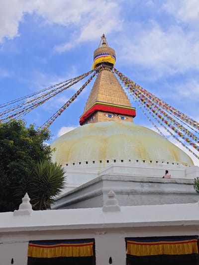 The image shows the Boudhanath Stupa, a significant Buddhist pilgrimage site located in Gokarneshwar, Nepal. The stupa is a large, whitewashed dome topped with a golden spire. Prayer flags in various colors are strung across the sky, adding to the spiritual atmosphere. The eyes of Buddha are painted on the base of the spire, a common feature of Tibetan Buddhist stupas. A single person, wearing a pink top, is seated on the edge of the stupa's circular walkway, looking out at the surroundings. Lush green trees frame the left side of the image, providing a natural contrast to the architectural grandeur of the stupa. The sky is a vibrant blue with scattered white clouds, suggesting a pleasant day. The foreground features a white decorative wall with smaller stupa-like structures and colorful banners hung over openings.