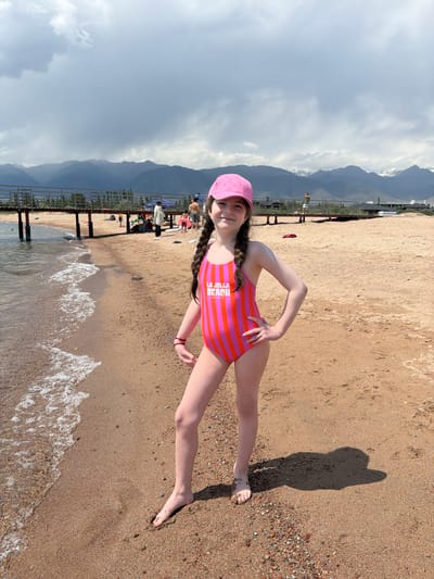 A young girl, wearing a pink and orange striped swimsuit and a pink cap, stands on a sandy beach. She is looking at the camera with a slight smile, her hand on her hip. Her hair is braided into two pigtails. She is positioned in the foreground, with waves lapping at the shore behind her. In the background, there is a pier with several people on it, and further back, mountains under a cloudy sky. The scene appears to be set in the afternoon, with the weather being partly cloudy. The text "LA JOLLA BEACH" is visible on her swimsuit. The location is Kara-Oy, Kyrgyzstan.
