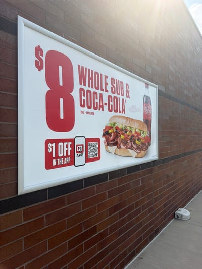 An outdoor advertisement is mounted on a brick wall. The advertisement is a large rectangular sign with a white background and red lettering. It promotes a "Whole Sub & Coca-Cola" deal for $8. A graphic of a large, filled sub sandwich is on the right side of the sign, next to a bottle of Coca-Cola. Below the deal information is a section that says "$1 OFF IN THE APP" with a QT app logo and a QR code. The sign is illuminated by sunlight from the upper right. The brick wall is made of reddish-brown bricks with dark mortar. The ground in front of the wall is a concrete sidewalk. A small, white robotic device is on the sidewalk to the right of the sign. The scene appears to be a commercial area.