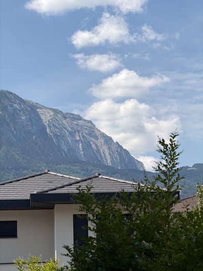 The image, oriented vertically, captures a serene mountain landscape. A large, rocky mountain dominates the center, its slopes covered in a sparse layer of vegetation. The sky above is a clear blue, dotted with scattered white clouds. In the foreground, the roof of a modern building, with dark grey tiles and white walls, is visible. Some of the building's windows, with dark shutters, are also present. A branch of a leafy green tree extends into the frame from the top right corner, adding a touch of nature to the architectural element. The overall impression is one of a peaceful, possibly rural setting, with the majestic mountain providing a dramatic backdrop to the man-made structure. The lighting suggests it is daytime, likely morning or afternoon, with bright sunlight illuminating the scene. No people or explicit text are visible in the image. The location context provided is Cluses, France.