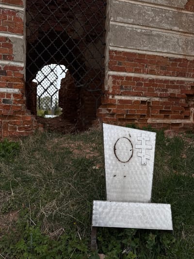 The image displays a scene in Staroe Rakomo, Russia, featuring a combination of an old, dilapidated red brick structure and a metal sign resting on grassy ground.
The primary subject on the left is a crumbling brick wall, characteristic of an ancient or ruined building. The red bricks are visible, with sections of deteriorating mortar and what appear to be older concrete repairs or decorative strips running vertically. Large openings, possibly windows or doorways, are present in the brickwork. One prominent opening in the foreground is partially covered by a dark, patterned metal mesh or grille, through which a brighter, indistinct area is visible beyond.
To the right, a silver-colored metal sign lies on the ground amidst wild grass and weeds. The sign is rectangular and features two distinct shapes cut out or etched into its surface. Near the top, a horizontal pattern resembling interconnected hash symbols or a stylized fence is present. Below this, a large, empty oval shape is visible, containing a few small holes. The surface of the metal sign has a brushed or swirled texture.
The ground around the sign and the base of the brick structure is covered with a mix of green and dry grass, along with various leafy weeds. The lighting suggests a daytime setting, possibly overcast, as there are no strong shadows. The scene evokes a sense of decay and history, with the old brickwork slowly succumbing to time and nature. There are no people visible, and no discernible text appears on the sign or elsewhere in the image.
