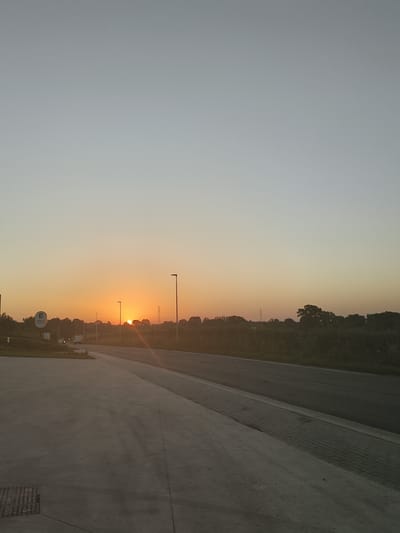 The image depicts an outdoor scene at dawn or dusk, with the sun low on the horizon. The sky transitions from a light blue or grey at the top to a warm orange and yellow hue near the horizon, indicating sunrise or sunset. In the distance, a line of trees and vegetation forms a natural border. Several streetlights are visible, with one prominently placed near the center. A road, appearing to be asphalt, curves through the scene, bordered by a paved walkway. On the left side of the image, a concrete area is visible, featuring a metal grate. A circular sign with text and a logo is also present on the left, partially obscured by vegetation. Several vehicles, including a white van and a car, are moving or parked along the road. The overall environment suggests a semi-rural or suburban setting, potentially near a road or a commercial area. The provided location of Thimister, Belgium, aligns with a possible European landscape.