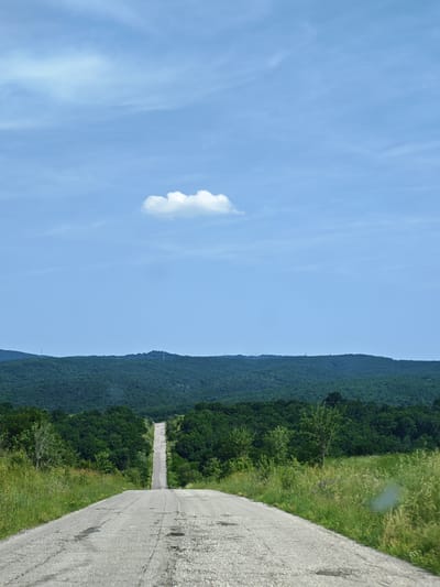 This image depicts a rural landscape near Dabovets, Bulgaria, under a clear, bright blue sky with scattered white cumulus and cirrus clouds. A long, narrow, asphalt road dominates the foreground, descending steeply into the distance. The road surface is visibly weathered with numerous cracks and potholes. Flanking the road are overgrown verges of tall, green, and dried grass, along with some small deciduous trees and bushes. In the midground, dense, dark green deciduous forest covers undulating terrain. The horizon is defined by a series of forested hills, above which the sky extends. Several faint, vertical structures, possibly power line towers or communication masts, are discernible along the crests of these distant hills. No people, vehicles, or animals are visible in the scene. The overall environment suggests a summer day in a remote, natural setting.