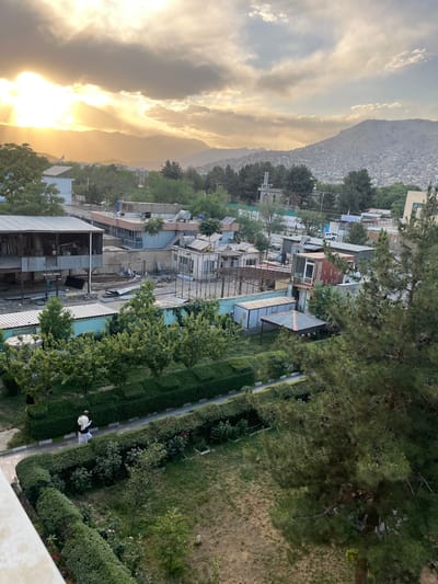 The image captures a panoramic view of Kabul, Afghanistan, during sunset. The sky is a dramatic mix of bright sunlight filtering through thick clouds, casting an orange glow over the landscape. In the foreground, a residential area is visible, with a variety of buildings ranging from unfinished structures to more developed houses. There are numerous trees and manicured hedges, suggesting a well-maintained area within the city. A person dressed in traditional clothing, a white tunic and dark pants, walks along a pathway bordered by hedges. In the background, the city sprawls across the hills, with mountains forming a dramatic backdrop. The overall scene depicts a blend of urban development, natural elements, and a sense of evening tranquility.