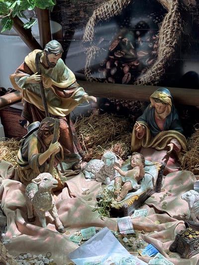 A nativity scene displays figurines of Mary, Joseph, and baby Jesus in a manger surrounded by hay. Shepherds and sheep are also present. Coins, paper money, and a drawing are scattered around the scene. A backdrop depicts a coffee bean bag.