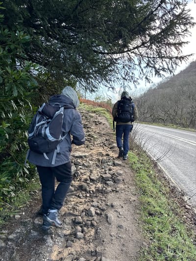 Two individuals are depicted walking uphill on a rocky dirt path in High Peak, United Kingdom. The person in the foreground wears a grey hooded jacket, black trousers, and hiking boots, carrying a black and grey backpack. Ahead, a second individual wears a green hooded jacket, blue jeans, and dark hiking boots, with a black backpack. The path runs parallel to an asphalt road on the right, which features a double white line and is bordered by sparse green grass and bare bushes. To the left, dark green evergreen shrubs and a large conifer tree with dense foliage overhang the path. In the background, a hillside covered with leafless deciduous trees ascends under a light blue sky.