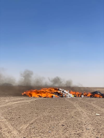 In Daman, Afghanistan, a large pile of waste is ablaze in a barren, arid landscape. The fire produces thick plumes of dark gray smoke that billow upwards and spread across the horizon. The flames are intense, with bright orange and yellow visible. The waste appears to consist of white boxes, some of which are intact and stacked, while others are burning and disintegrating. A small amount of yellow material is also visible within the burning debris. The ground surrounding the fire is dry and dusty, with tire tracks suggesting vehicular activity. The sky above is a clear, pale blue.