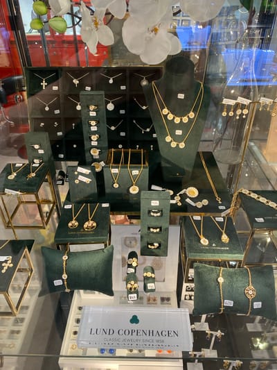 This image shows a display of gold jewelry, primarily necklaces and earrings, in a storefront window. The jewelry is arranged on dark green velvet stands and within glass cases. Several items feature small, white, daisy-like charms with yellow centers. Prices are visible on some of the items, such as "1250," "975.00," and "895." The display includes the branding "LUND COPENHAGEN CLASSIC JEWELRY SINCE 1858." The background shows a blurred red cabinet and reflections in the glass. This is a storefront display, likely for Lund Copenhagen, located in the city of Herning, Denmark.