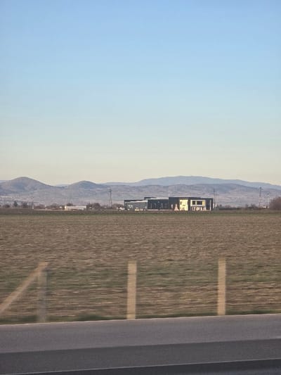 The image displays a panoramic view of a rural landscape adjacent to Diadovo, Bulgaria. The foreground is defined by a dark grey asphalt surface, likely a road, with faint, vertical, light-colored reflections suggesting it was captured from within a vehicle. Immediately beyond the road, a vast, flat agricultural field occupies the midground, characterized by brown soil and sparse patches of green vegetation. This field exhibits horizontal motion blur, consistent with movement during capture. In the mid-distance, a prominent, dark-toned industrial or commercial building with a flat roof is centrally situated. This structure features a dark facade with distinct rectangular lighter-colored sections on its right side. Surrounding this building, additional smaller structures and multiple utility pylons are visible. The background is composed of a continuous range of low, hazy mountains or hills stretching across the horizon. The sky above is clear, transitioning from a light blue at the zenith to a paler hue closer to the horizon, encompassing the upper half of the frame. No individuals or specific actions are discernible within the scene.