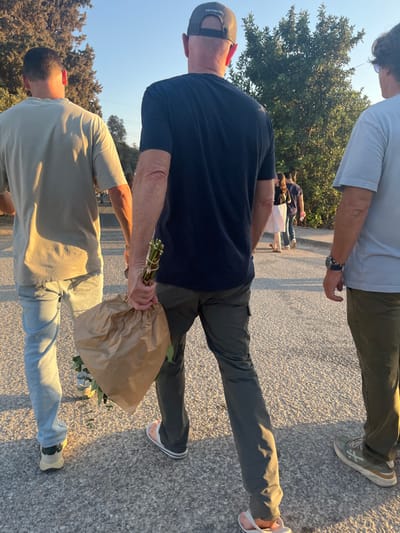 The image captures three adult males walking away from the camera on a paved surface during late afternoon or early evening in the Brener Regional Council, Israel. The man on the left wears a light beige short-sleeved t-shirt, light blue jeans, and dark-soled sneakers. The central figure wears a dark blue short-sleeved t-shirt, dark olive-grey cargo pants, white flip-flops, and a grey mesh baseball cap with a visible "Carhartt" logo. He holds a bundle of long-stemmed plants, partially wrapped in crumpled brown paper, in his right hand. The man on the right wears a light blue short-sleeved t-shirt, dark olive-khaki pants, and light-colored sneakers; a dark wristwatch and a beaded bracelet are visible on his left wrist. All three are moving forward. Further down the path, several other individuals are visible walking, including one wearing a white skirt. The environment features green trees and foliage lining the path under a clear blue sky, illuminated by strong, low-angle sunlight casting long shadows.