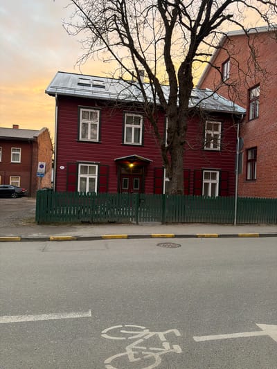 The image depicts a street scene in Tartu, Estonia. A two-story red wooden house with white-framed windows stands behind a dark green wooden fence. A bare tree partially obscures the view of the house. Adjacent to the red house is a brick building. The sky shows a sunset glow. A street with a designated bicycle lane marked with a bicycle symbol is visible. A dark-colored car is parked on the street near a sign that is partially visible.