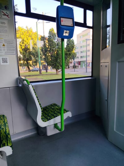 The image depicts the interior of a public transportation vehicle, likely a tram or bus, in Arad, Romania. The focus is on an empty seat with green and black patterned upholstery, a green pole extending from the floor to a blue display unit mounted on the window, and the window itself revealing an urban street scene. Outside the window, there are trees with yellowing leaves, a building with multiple stories and balconies, and a street with a parked car and traffic lights. The interior of the vehicle is clean and modern, with a grey floor and white walls. A digital display unit on the window shows route information and a Romanian flag, confirming the location. No people are visible in the image. The overall impression is that of a quiet, daytime scene inside a public transport vehicle.