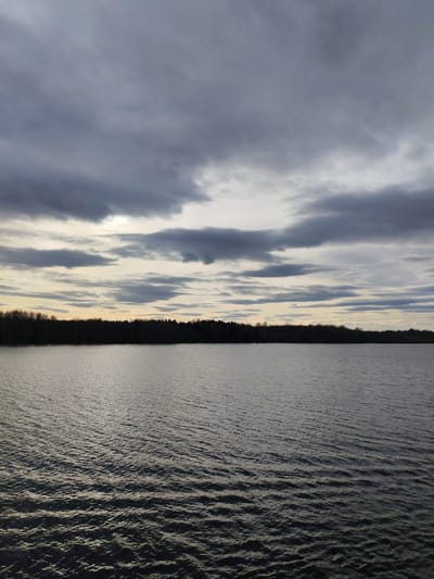 The image depicts a body of water, possibly a lake or a wide river, under a cloudy sky. The water is rippled, indicating a slight breeze. A dark treeline is visible in the distance, separating the water from the sky. The sky is filled with grey and white clouds. The location is possibly in the city of Yakunina Gora, Russia. There are no people visible.