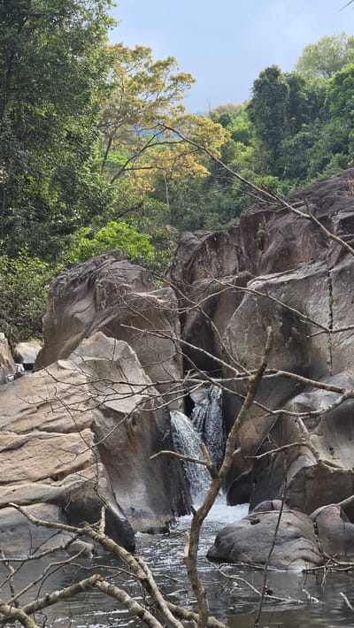 The image shows a rocky waterfall in a natural setting. Gray and brown rocks form the landscape, with sparse, leafless branches in the foreground. Lush green trees and foliage are visible behind the rocks. A small portion of the waterfall can be seen flowing through the rocks. The image is taken in the city of Nam Ninh Hoa, Vietnam.