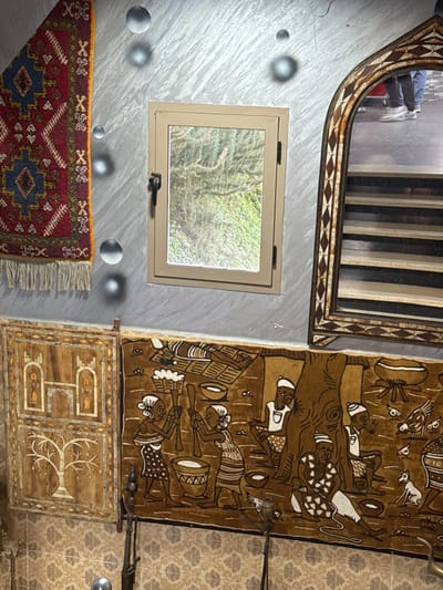 This image depicts an interior wall space within a structure located in Zerkten, Morocco. The upper portion of the wall is textured, appearing light gray with multiple embedded, reflective spherical elements. A central, rectangular window with a light brown frame and a black latch on the left provides a direct view of dense green foliage outside.
To the left of the window, an ornamental textile, predominantly red, blue, and cream with geometric patterns and fringe, is diagonally draped. Below this and to the lower left, a rectangular wooden panel, adorned with light-colored inlay depicting a stylized building and a tree, hangs vertically.
To the right of the window, an arched mirror with an elaborately carved wooden frame, featuring geometric inlay patterns, is mounted. The mirror reflects a portion of a staircase with horizontal risers and the lower legs and feet of two individuals wearing shoes, one white sneaker and one dark shoe, positioned on a landing above.
The lower portion of the wall is covered with patterned tiles or wallpaper. Below the window and extending to the right, a large brown tapestry or textile art piece is displayed. This artwork features a detailed scene in lighter brown outlines, depicting multiple figures engaged in activities such as grinding with pestles and mortars, seated individuals, a recumbent figure, a tree, birds, and a dog.
Partially visible at the bottom of the image are sections of a patterned floor and dark, metallic stand-like objects. No individuals are directly present within the room shown, but human presence is indicated by the mirror's reflection.