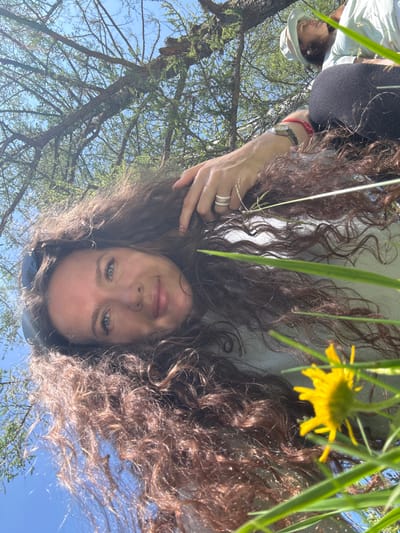 A woman with long, curly brown hair is lying on her back outdoors, with her head tilted to the left. She is looking towards the camera with a slight smile. Her left hand is resting on her head, and she is wearing a watch with a red band on her left wrist, and two rings on her left index finger. In the background, there are tree branches and a clear blue sky. To the right of the woman, another person is partially visible, wearing a light-colored hat and shirt. A yellow flower and some green grass are in the foreground, obscuring some of the woman's body and hair. The image appears to be taken from a low angle, looking up at the woman and the trees.
