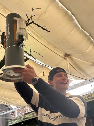 The image shows a young man wearing a black baseball cap and a navy and white striped rugby shirt, smiling while holding a beer dispenser. The dispenser, a dark gray cylindrical device with a clear plastic cup holder attached, is positioned above his head. The man's right arm is extended upwards, holding the base of the dispenser. A silver wristwatch is visible on his left wrist.

The background features a textured, light-colored canopy, likely made of fabric, stretched overhead. Bare branches of a tree are visible against the canopy. In the lower portion of the image, a building with a reddish roof and a green sign with text is visible. The sign appears to be for "VILLAGE PE" and "TO NSBERG" with additional text, suggesting the location is Tønsberg, Norway. The overall lighting suggests an outdoor setting during the daytime. The man's engagement with the beer dispenser and his happy expression imply an activity related to serving or receiving a drink.