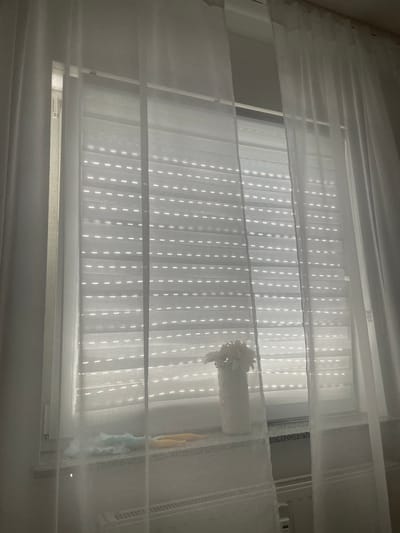 The image shows a window with white sheer curtains in front of vertical blinds. The blinds have a pattern of small, evenly spaced vertical dashes of light shining through them. A white vase with a white flower decoration sits on the windowsill to the right of the blinds. The scene is dimly lit, suggesting it is either early morning or late evening. The sheer curtains are draped and layered, adding a soft, diffused quality to the light. The overall impression is one of a quiet, indoor domestic setting. There is no visible text. The location context is Wiehl, Germany.