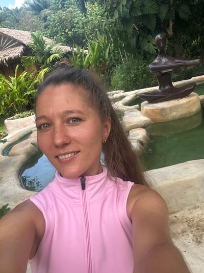 A woman with blonde hair tied in a ponytail and green eyes is taking a selfie in front of a pond with a statue of a person meditating in the water. She is wearing a pink sleeveless top with a zipper. The background features lush green foliage and a thatched-roof building, suggesting an outdoor, tropical location. The scene appears to be in Tambon Khlong Thom Nuea, Thailand.