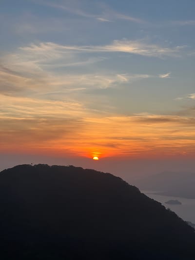 This is a vertical, landscape photograph capturing a sunset over a mountainous region, likely El Cacao, El Salvador. The sky is ablaze with color, transitioning from a pale blue at the top left to vibrant oranges and reds towards the center. Wispy clouds are scattered throughout, catching the sunlight and adding texture to the sky.

The sun is partially obscured by a dark silhouette of a mountain range, which dominates the right side of the frame. The mountain's jagged edge creates a stark contrast against the colorful sky. In the distance, other misty, mountainous shapes are visible, hinting at a vast and rugged landscape.

The time of day is clearly sunset, as indicated by the intense colors in the sky and the low angle of the sun. The weather appears clear, allowing for a dramatic sunset display. The location context suggests a rural or mountainous area in El Salvador. There are no people or man-made objects explicitly visible, contributing to a sense of natural beauty and tranquility. The overall mood evoked is one of awe and peace, typical of a stunning sunset. There is no visible text in the image.