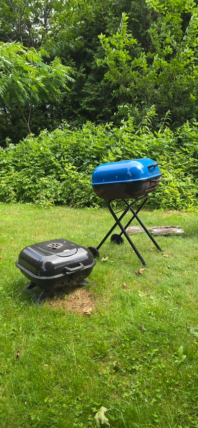 The image shows two charcoal grills in an outdoor setting. One grill, a blue and black kettle-style grill on a black folding stand, is positioned on the left side of the frame. The other grill, a black portable grill, is located on the right. Both are situated on a lush green lawn. In the background, dense green foliage from trees and bushes forms a natural border. The scene appears to be a backyard or park setting. The lighting suggests it is daytime, with the sun casting reflections on the blue grill lid. There are no people visible, and the grills are not in use, indicating a static scene rather than an active event. The overall impression is one of a calm, outdoor environment, possibly during summer in Drummondville, Canada.