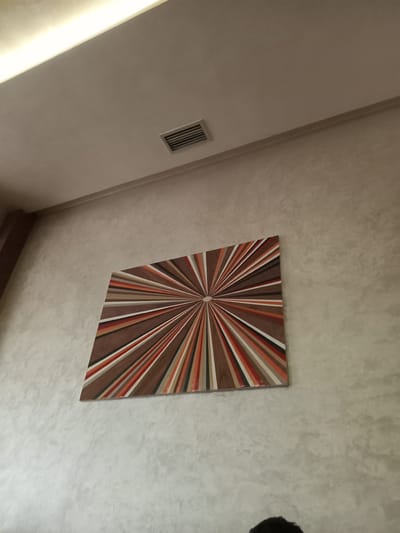 The image presents an indoor scene, viewed from a low angle looking upwards towards a ceiling. The ceiling is painted in a light, textured off-white color, possibly stucco or a similar finish. A square artwork featuring a radiating pattern in shades of brown, orange, and white is mounted on the ceiling. To the left of the artwork, there is a rectangular air vent also on the ceiling. A dark, out-of-focus object is visible in the upper right corner of the image, possibly part of a light fixture or another ceiling-mounted item. Portions of walls are visible on the left and bottom of the frame, painted in a lighter off-white hue, providing depth to the image. The image does not provide conclusive visual elements related to Ksar-el-Kebir, Morocco.