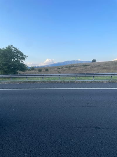 The scene depicts a rural landscape located in the outskirts of Paliano, Italy, under a clear blue sky. In the immediate foreground, a dark asphalt road surface occupies the lower half of the image, featuring a continuous white demarcation line along its upper edge. Above the road, a segmented, galvanized steel guardrail extends horizontally across the frame, positioned at the base of a grassy and sparsely vegetated embankment. To the left, a large, dense deciduous tree with dark green foliage rises from behind the guardrail. Further right, atop a gently sloping, dry, golden-brown hill, a smaller, solitary dark green tree is visible. In the distant background, undulating terrain transitions into a faint blue mountain range. Several distinct white cumulus clouds are scattered near the mountain peaks. No individuals, vehicles, or animals are present. The lighting suggests daytime conditions with ample natural light.