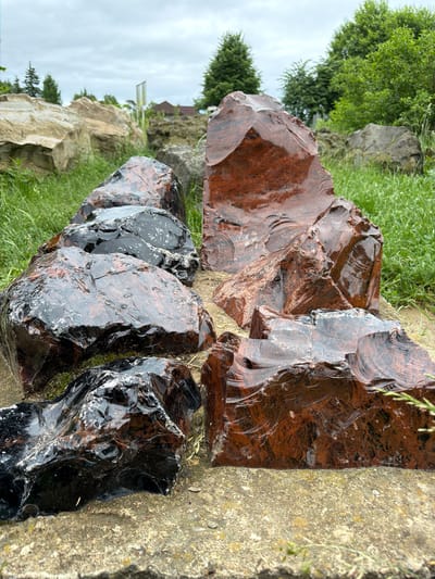 This media file shows a close-up, angled view of several large, rough-hewn pieces of obsidian, a volcanic glass. The obsidian is displayed in an outdoor setting. Some pieces are a deep, glossy black, while others have a rich reddish-brown or mahogany hue, with some showing streaks and inclusions of lighter colors. The surfaces of the obsidian are highly reflective, catching the light and revealing their sharp, conchoidal fracture patterns.
The obsidian pieces are resting on a textured, concrete-like surface, with blades of green grass growing in between and around them. In the background, more natural rock formations are visible, interspersed with lush green grass and trees. The trees appear to be deciduous and are in full leaf, suggesting a warmer season, likely spring or summer. The sky is overcast, indicated by the diffused light and muted colors, suggesting a cloudy day. The overall impression is one of a natural or landscaped garden setting, possibly designed to showcase these striking geological specimens. No people are visible, and no text is present in the image. The scene evokes a sense of raw natural beauty and geological wonder.