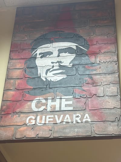 The image shows a large, framed piece of art, possibly a mural or a stretched canvas, depicting a stylized portrait of Che Guevara. The artwork is set against a background that mimics a brick wall, with red and grey bricks. Che Guevara's face is rendered in black, white, and grey tones. Below the portrait, the words "CHE GUEVARA" are printed in large white sans-serif font, also designed to appear as if painted on the brick surface. The artwork is mounted on a wall, with the top edge of the frame visible against a textured ceiling. The bottom portion of the image shows a beige-colored wall below the artwork. There are no people visible in the image. The context of Maloyaroslavets, Russia, is not visually supported by the image content.