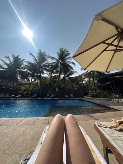 A bright, sunny scene depicts a poolside setting. Turquoise water stretches alongside a stone tile patio where a sunbather reclines on a striped towel. The person's wet legs glisten in the light. Palm trees sway in the background, partially shaded by an open white parasol. The sun, a radiant burst of light, illuminates the serene scene.