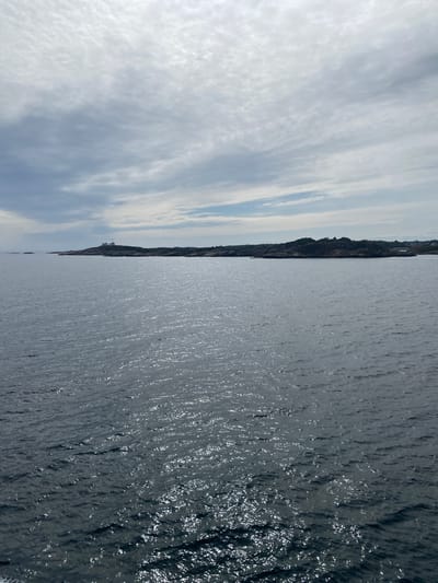 The image shows a vast expanse of dark, choppy water in the foreground, with sunlight reflecting off the surface in shimmering patterns. In the distance, a rocky, sparsely vegetated coastline stretches across the horizon. On the furthest point of the landmass, a white lighthouse with a red roof is visible. The sky above is overcast with a mottled pattern of grey and white clouds. The scene depicts a coastal landscape, likely near Stavern, Norway, characterized by its rugged terrain and the presence of a lighthouse. There are no people or specific actions depicted in the image.