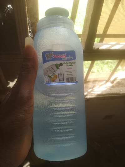 A light blue, textured plastic water bottle is held in a person's hand. The bottle has a blue and white label with text and an illustration of water pouring into a glass, along with lemons. The label reads "SACVIN Cold Water Bottle 1000ML INDOOR & OUTDOOR LEAK PROOF RECYCLABLE FREEZER SAFE". The background shows a wooden structure with netting and foliage, suggesting an outdoor or semi-outdoor setting. The lighting suggests it is daytime. The scene is not specifically identifiable as being in Majemu, Nigeria; it is a generic image of a water bottle.