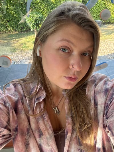 An image captured in San Phra Net, Thailand, depicts a female individual taking a selfie indoors. The subject has long, light brown hair, light blue eyes, and a small silver hoop nose ring on her right nostril. A white wireless earbud is visible in her right ear. She wears a light pink and purple plaid button-up shirt over a solid light brown top, accessorized with a silver-colored necklace featuring an angular, possibly 'K'-shaped pendant. The background behind the individual features a large window showcasing an outdoor area with dense green foliage and a light-colored ground surface. Blurred, abstract structures with visible orange and blue accents are partially visible through the window. Natural light illuminates the scene.