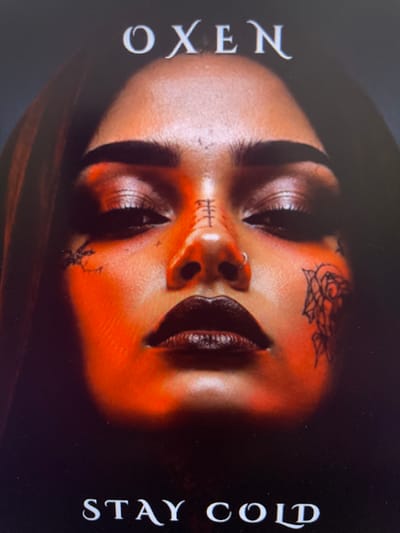 The media file features a close-up, upside-down portrait of a person's face. The subject has dark lipstick and a nose ring. There are several tattoos visible on their face, including a rose tattoo on the forehead and intricate designs around the mouth and chin. The lighting is dramatic, casting strong shadows and highlighting the contours of the face with an orange-toned glow.
The words "OXEN" are displayed vertically on the left side of the image in a stylized font. On the right side, also displayed vertically, are the words "STAY COLD" in a similar font.
The image appears to be a cover or promotional material, possibly for music or a creative project. There are no explicit environmental cues suggesting a specific time of day, weather, or location beyond the provided context of Punta Ala, Italy, which is not visually evident in the image itself. The overall mood conveyed is intense and possibly edgy, with the tattoos and dark makeup contributing to a stylized aesthetic.