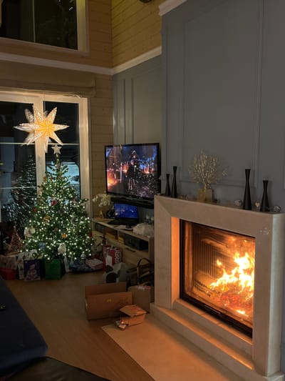 A cozy living room features a fireplace with blazing flames, a television displaying a film, and a decorated Christmas tree surrounded by presents. Light streams through a window adorned with a large star. The room is decorated with Christmas ornaments and festive decorations. A couch sits in the lower right corner. The style could be considered modern with neutral colors.