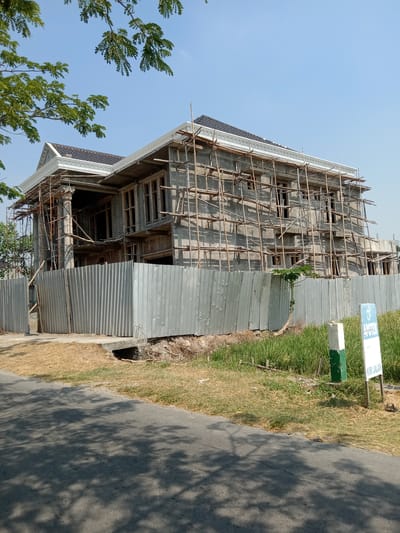 A two-story house under construction in Kemamang, Indonesia, is surrounded by scaffolding made of bamboo poles. The house's exterior is unfinished concrete, with visible window frames. A corrugated metal fence encloses the property, and a road with tree shadows stretches across the foreground. A signpost stands near the fence, indicating directions. The sky is clear and blue, with some tree branches in the upper left corner. The environment appears dry, with patches of grass and straw around the construction site.