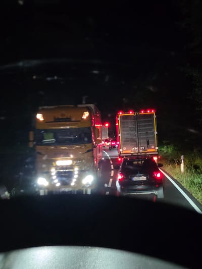 The image depicts a traffic jam on a road at night, likely in the vicinity of Seligenstadt, Germany. A yellow truck with its headlights on is prominently in the foreground, facing away from the viewer. Behind it, a black car with its brake lights illuminated is visible, displaying a license plate reading "AB·RD 6666". Further ahead, another truck with a dark-colored trailer is stopped, its red taillights a bright contrast to the darkness. The road surface is visible, with white lane markings. The surrounding environment is dark, with hints of greenery and foliage on the right side of the road, suggesting a rural or semi-rural setting. The overall impression is one of a halted flow of traffic under the cover of night.