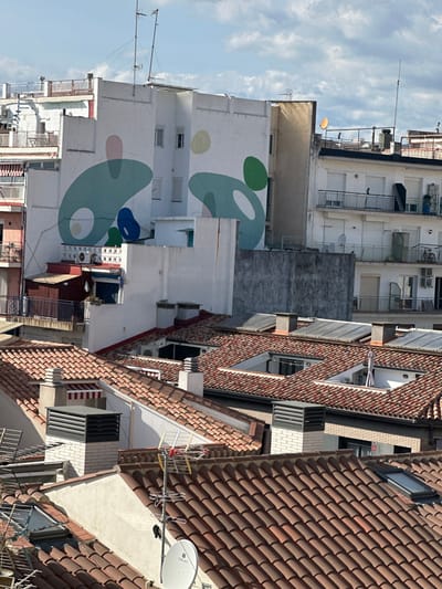 This image captures an urban landscape in Pineda de Mar, Spain, from an elevated perspective under a partly cloudy sky. The scene is dominated by a dense arrangement of residential buildings and their rooftops.

A prominent, multi-story white building in the midground features a large, abstract mural on its side facade. The mural consists of fluid, organic shapes in varying shades of teal, blue, green, light pink, and yellow. Below this building, a complex arrangement of lower white structures and a small, dark wooden shed with a blue door are visible, along with multiple air conditioning units.

In the foreground and midground, numerous terracotta-tiled rooftops stretch across the frame, showcasing various pitches and architectural elements. These roofs include multiple chimneys, some topped with black louvered vents, and several rectangular skylights. Satellite dishes, including one clearly marked "Triax," and television antennas are mounted on various rooftops. One rooftop appears to have a small terrace with a white patio umbrella visible.

Surrounding the central mural building are other multi-story residential buildings with balconies featuring white railings. One balcony displays a bicycle. Windows on these buildings are varied, some with closed white shutters. Multiple external air conditioning units are attached to facades. The overall environment suggests a densely populated residential area with typical Mediterranean urban architecture. No individuals are visible, and no specific actions or interactions are depicted.