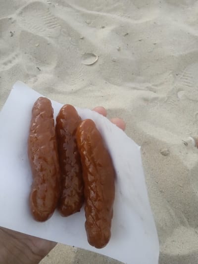 A partial human hand is depicted holding three elongated, brown, glazed food items, possibly sausages or fried pastries, wrapped in a crumpled piece of white paper. The background consists of light-colored sand, showing faint footprints, scattered small shells, and minor debris, indicating a beach or dune environment. This scene is located in Al-Mawasi, Palestinian Territories.